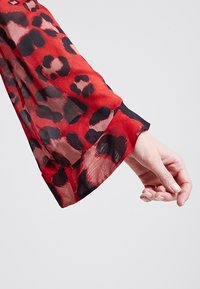 Blusa rossa trasparente con maniche, caratterizzata da un motivo leopardato nero e rosso. Il tessuto sembra leggero con un design fluido.
