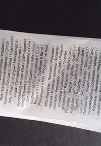 Valkoinen tekstiilien hoito-ohje -label, jossa on useita kieliä; luettelee materiaalit, mukaan lukien 95 % kierrätetty polyesteri ja hoito-ohjeet.