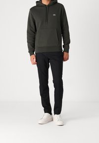 Felpa con cappuccio verde scuro con tasca frontale e coulisse, abbinata a pantaloni neri e sneakers bianche. Design semplice e casual.