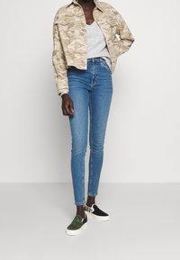 Mujer vestida con chaqueta de camuflaje beige, camiseta gris, jeans ajustados azules y zapatos de deslizar desiguales con patrones de camuflaje y cebra.