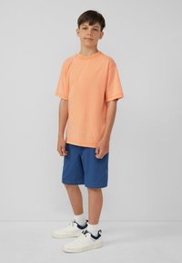 Oranje katoenen T-shirt met korte mouwen, gecombineerd met een blauwe short. Witte sneakers met marineblauwe accenten maken de outfit compleet. Eenvoudige en casual stijl.