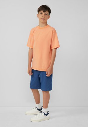 Oranje katoenen T-shirt met korte mouwen, gecombineerd met een blauwe short. Witte sneakers met marineblauwe accenten maken de outfit compleet. Eenvoudige en casual stijl.