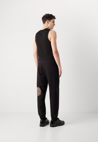 Black sleeveless tank top paired with black sweatpants featuring a textured patch on the right knee and elastic cuffs. Black sneakers complete the look.
