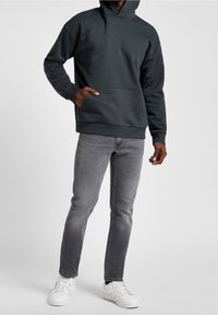 Hombre con sudadera gris oscura, jeans ajustados grises y zapatillas blancas, de pie contra un fondo claro liso.