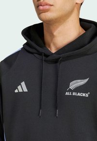 Sort hættetrøje med en front kangaroo lomme, snoretræk og grå "ALL BLACKS" logo med et bregne. Akcentstriber i lyseblå på ærmerne.