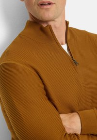 Brauner Reißverschluss-Pullover mit strukturiertem horizontalem Rippenmuster, hohem Kragen und engen Bündchen. Das Material wirkt weich und gestrickt.