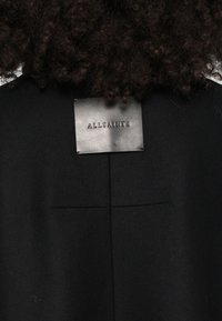 Μαύρο μάλλινο παλτό με υφή, διαθέτει ορθογώνια δερμάτινη ετικέτα με ανάγλυφο το λογότυπο "ALLSAINTS" σε ασημί. Απλός, ραμμένος σχεδιασμός.