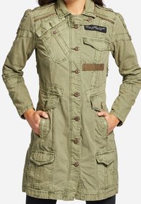 Olivgrön, militärinspirerad jacka med knappstängning, flera fickor och kontrasterande texturerade lappar; har en funktionell krage och dragkedjedetaljer.