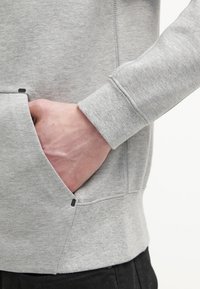 Hand rustend in de voorzak van een lichtgrijze sweatshirt met zichtbare stiksels op de mouw en de zijkant.
