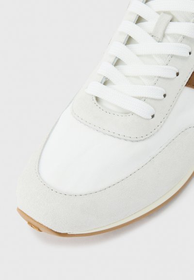 Κοντινό πλάνο δείχνει το μπροστινό μέρος ενός λευκού αθλητικού παπουτσιού με επένδυση από σουέντ, λευκά κορδόνια και ελαφρώς καφέ λαστιχένια σόλα σε απλό φόντο.