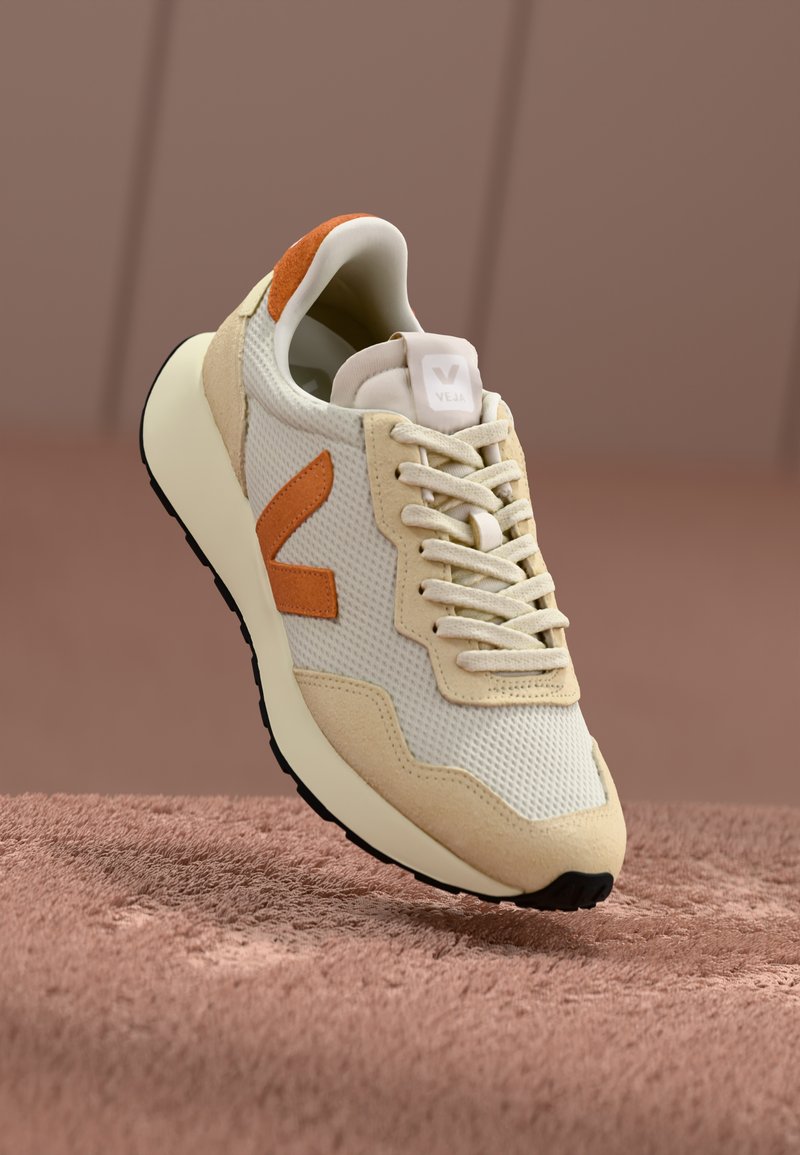 Beige sneakers med mesh- og semsket overdel, oransje aksent og hvit såle. Har snøring, logo på tungen og teksturert overflate.