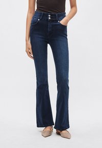 Jeans azul oscuro de talle alto con pierna acampanada, con dos bolsillos delanteros y detalles de botones plateados. Textura de mezclilla suave.