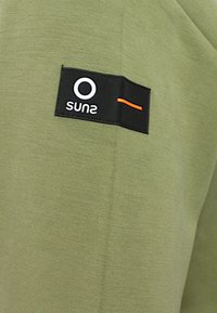 Tessuto verde oliva con una superficie liscia. Presenta un'etichetta nera con un logo circolare bianco e una linea arancione. Design semplice e moderno.