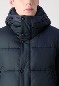 Veste doudoune marine avec col haut, fermetures à boutons et texture matelassée. Elle comprend des poches latérales et un design épuré.