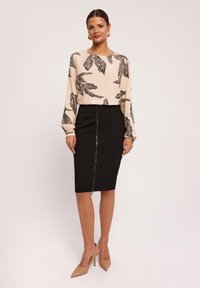 Beżowa bluzka z czarnym wzorem liści, długie rękawy, połączona z dopasowaną czarną spódnicą ołówkową z zamkiem z przodu. Beżowe buty na wysokim obcasie.