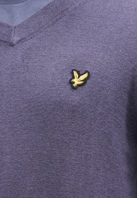 Maglione a scollo a V in tessuto viola morbido, con un piccolo logo di uccello ricamato in nero e oro sul lato sinistro del petto.