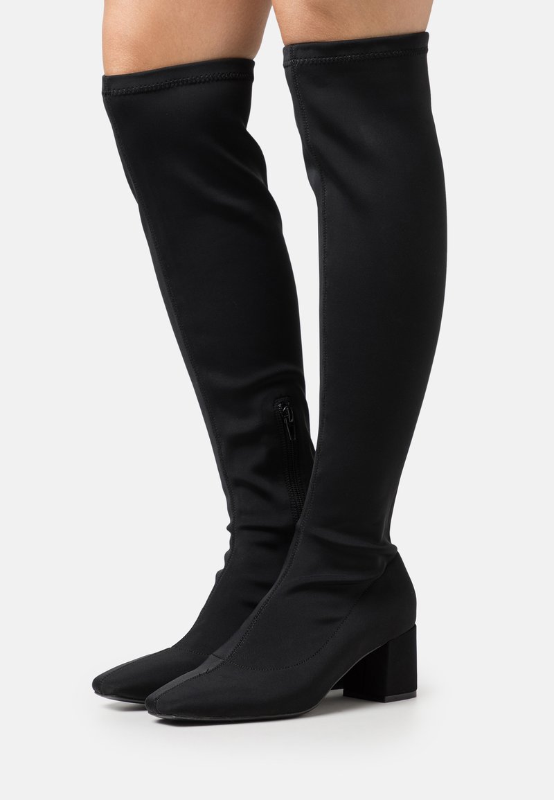 Botas negras por encima de la rodilla hechas de un tejido elástico. Presentan una punta afilada y un tacón cuadrado. Cierre lateral con cremallera para facilitar su uso.