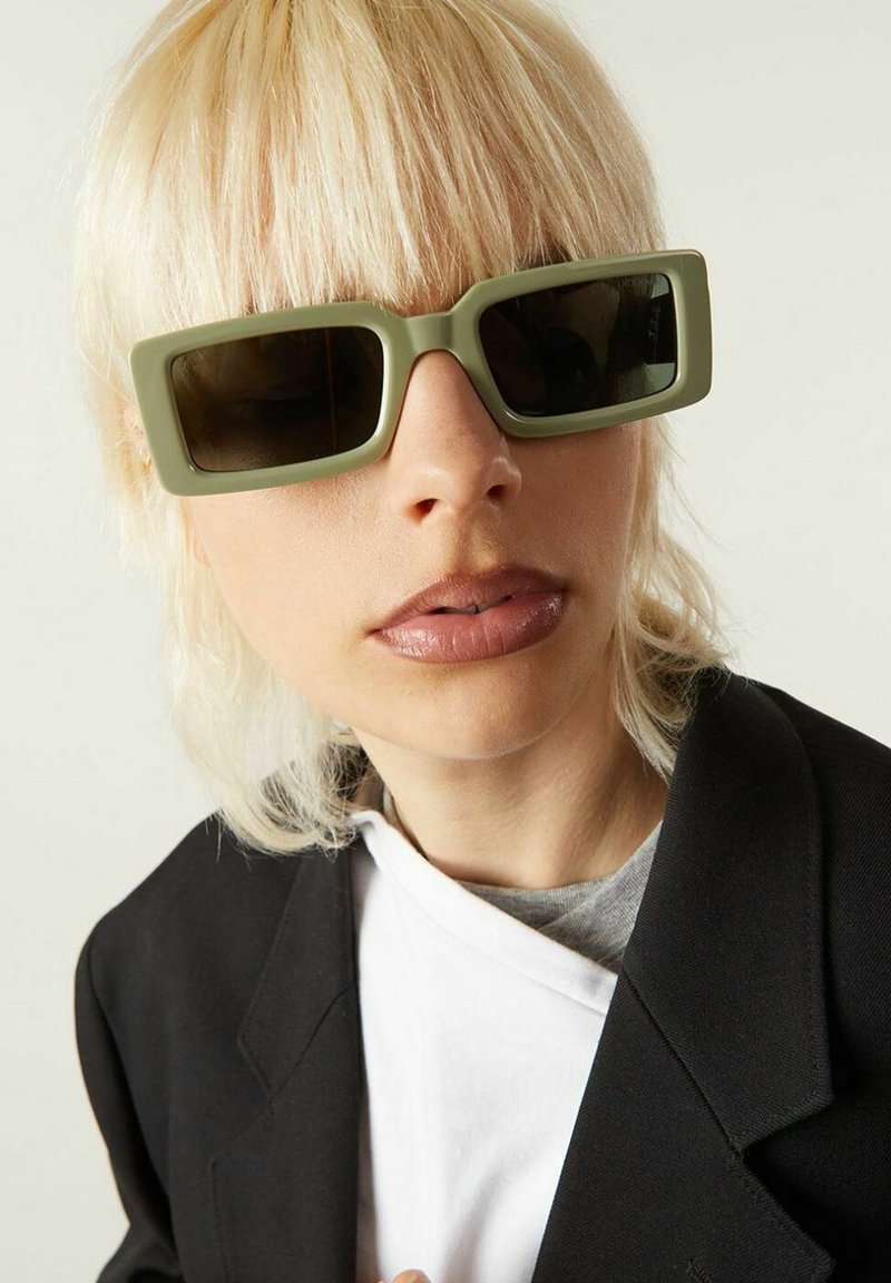 Lunettes de soleil vertes rectangulaires avec des verres sombres. La monture a une finition mate. La personne qui les porte a les cheveux courts, blond clair et porte un blazer noir.