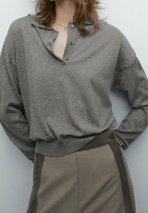Personne portant une chemise grise ample à manches longues avec un col partiellement déboutonné et un pantalon beige taille haute avec des bandes latérales foncées et un détail bouton.
