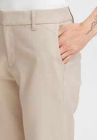 Pantalon beige avec une texture lisse, doté d'une taille ajustée et de poches latérales. Des coutures subtiles mettent en valeur le design ajusté.