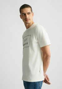 T-shirt bianca in cotone con scollatura rotonda e maniche corte, caratterizzata da un logo e testo stampati scuri sul frontale. Vestibilità casual.