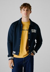 Marineblå varsityjakke med hvide stribede manchetter og krave, prydet med et logo på brystet, båret over en gul t-shirt og mørkeblå jeans.