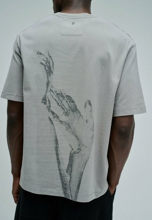 Homme portant un t-shirt gris clair avec une grande impression noir et blanc de deux mains qui s’étendent et se touchent dans le dos.