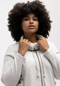 Chaqueta de cremallera gris claro con alto cuello, tejido texturizado, bolsillos frontales y detalles con cordón. Modelo sujetando el cuello.