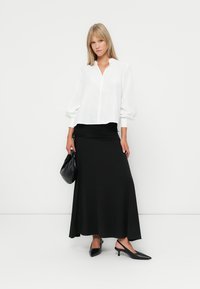 Witte blouse met knopen en volumineuze mouwen, gecombineerd met een zwarte, enkel-lange rok en zwarte hoge hakken. Zwarte handtas.