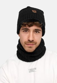 Black knitted beanie and matching neck warmer. Both feature ribbed texture and a small leather logo patch. Material appears soft and warm.