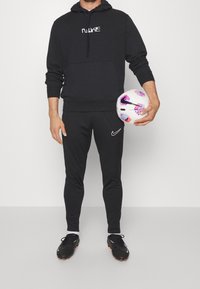 Musta huppari, harmaat verkkarit ja mustat nappikengät, kädessä kuvioitu jalkapallo, jossa on pinkkejä ja violetteja yksityiskohtia.