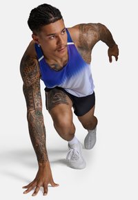 Sportowy top na ramiączkach przechodzący od niebieskiego do białego, zestawiony z czarnymi szortami. Widoczne tatuaże na ramionach i białe trampki ze sznurówkami.