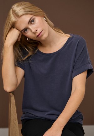 Blonde woman wearing a navy blue t-shirt, resting her head on her fist, seated on a wooden chair against a brown background.