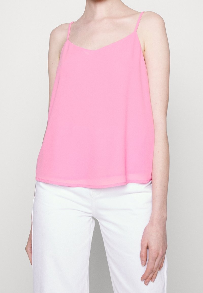 Femme portant un haut rose sans manches et un pantalon blanc, debout devant un fond clair uni.