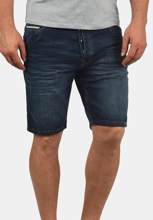 Mężczyzna ubrany w ciemnoniebieskie krótkie spodenki jeansowe i jasnoszarą koszulkę, stoi z rękami swobodnie przy bokach.