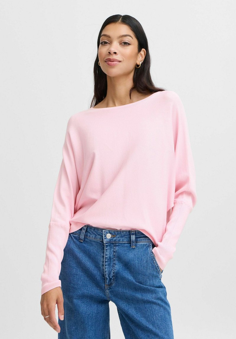 Sweater rose clair à manches longues avec un large décolleté bateau, associé à un jean en denim bleu. Texture douce et coupe décontractée.