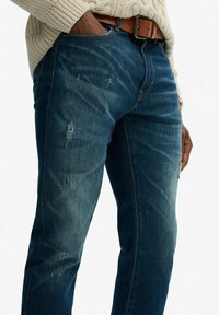 Persona vestida con jeans desgastados de color azul oscuro, cinturón marrón y un suéter de punto en color crema, con la mano reposando a un lado.