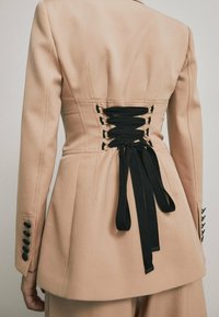 Blazer beige su misura con dettaglio di un nastro nero attillato sul retro, dotato di bottoni neri sui polsini e una vestibilità strutturata.