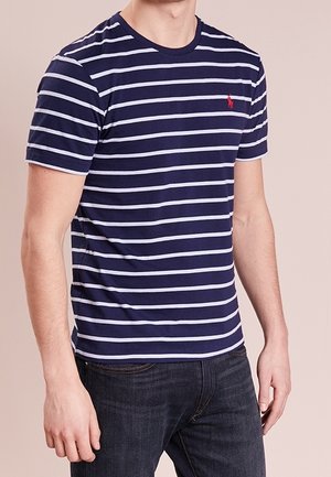 T-shirt à rayures navy et blanches à manches courtes avec col rond et un petit logo rouge sur la poitrine, associé à un jean foncé.
