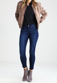 Femme portant une veste bomber taupe, une chemise noire boutonnée, un jean skinny bleu foncé et des bottines en daim grises, se tenant les mains dans les poches.