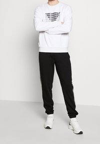 Sudadera blanca con estampado negro del logo "KARL LAGERFELD", combinada con pantalones de chándal negros ajustados con franjas laterales. Completo con zapatillas blancas.
