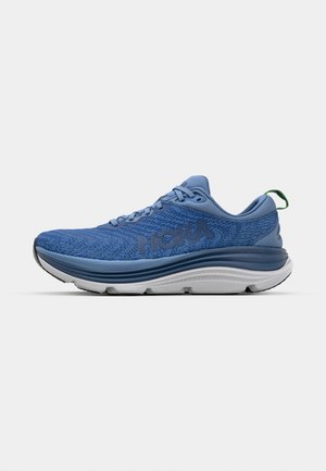 Chaussure de sport bleue avec un dessus texturé, dotée d'une semelle intermédiaire blanche, d'une semelle extérieure grise et d'une languette verte au niveau du talon. Logo HOKA visible.