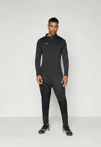 Svart treningsdress med lange ermer og halvlukket krage. Har et tonalt mønster på ermene og en kontrastfarget blå logo.