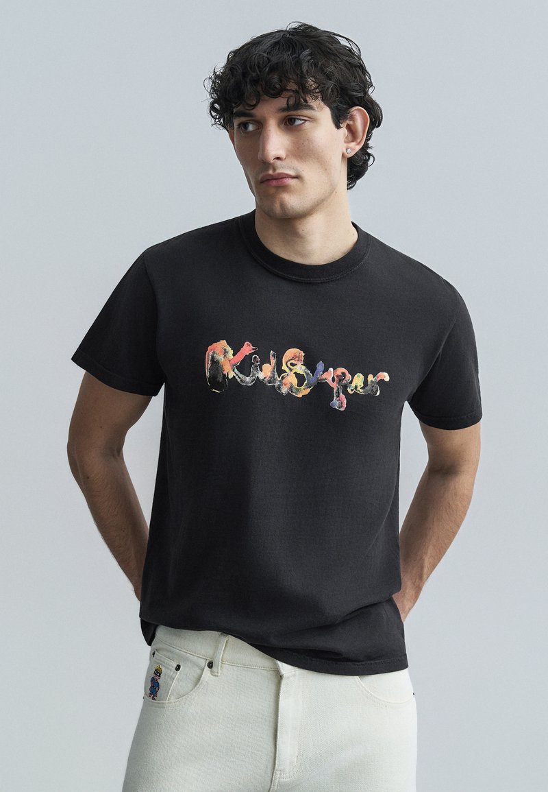 Μαύρο βαμβακερό T-shirt με πολύχρωμο γραφικό σχέδιο στο μπροστινό μέρος. Με κοντά μανίκια, λαιμόκοψη σε στυλ crew, φορεμένο με λευκό τζιν παντελόνι.