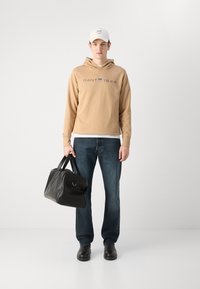 Felpa beige con logo "GANT 1949", jeans blu, borsa in pelle nera e scarpe nere; un cappellino da baseball bianco completa l'outfit.