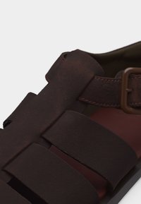 Κοντινό πλάνο σκούρου καφέ δερμάτινου σανδαλιού με φαρδιές λουρίδες και ρυθμιζόμενο αγκράφα στο πλάι.