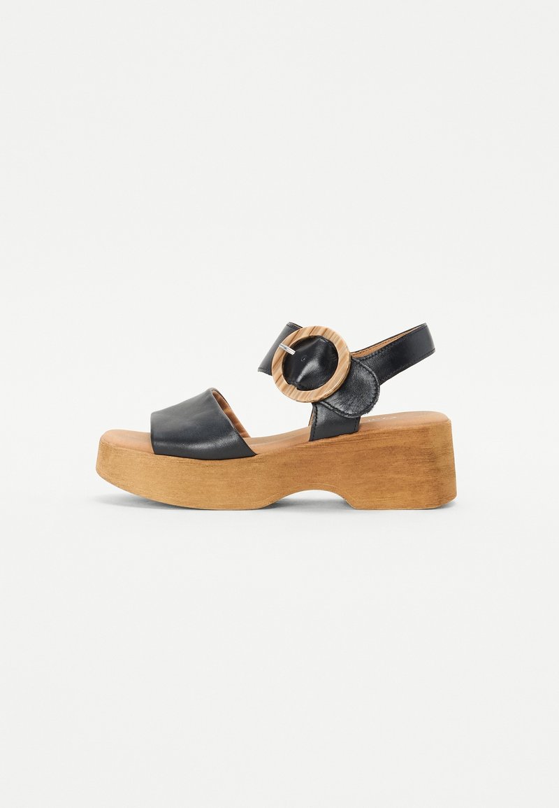 Sandale plateforme en cuir noir avec semelle en bois et grande boucle ronde en bois sur la bride de cheville, vue de côté sur fond blanc.