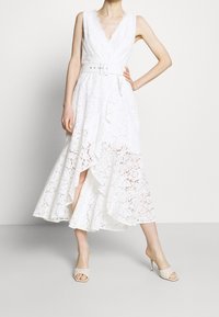 Robe blanche en dentelle avec un décolleté en V plongeant, sans manches, ourlet à volants et une large ceinture blanche. Portée avec des chaussures à talons nus et à lanières.