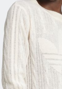 Suéter blanco de punto con un patrón texturizado, que presenta rayas verticales y un detalle de logo prominente. Material suave y ligero.