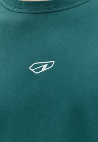 Tissu bleu sarcelle avec un petit logo blanc stylisé "N" à l'intérieur d'une forme trapézoïdale arrondie, centré près de l'encolure.